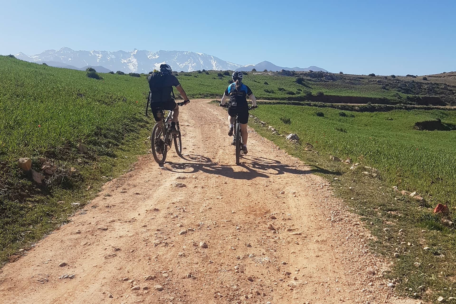 Two riders on a dirt track through green fields below the Atlas range
