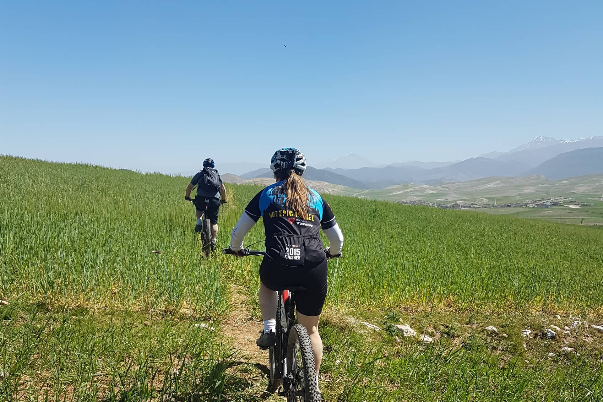 Two riders on a singletrack through green barley with the Atlas behind
