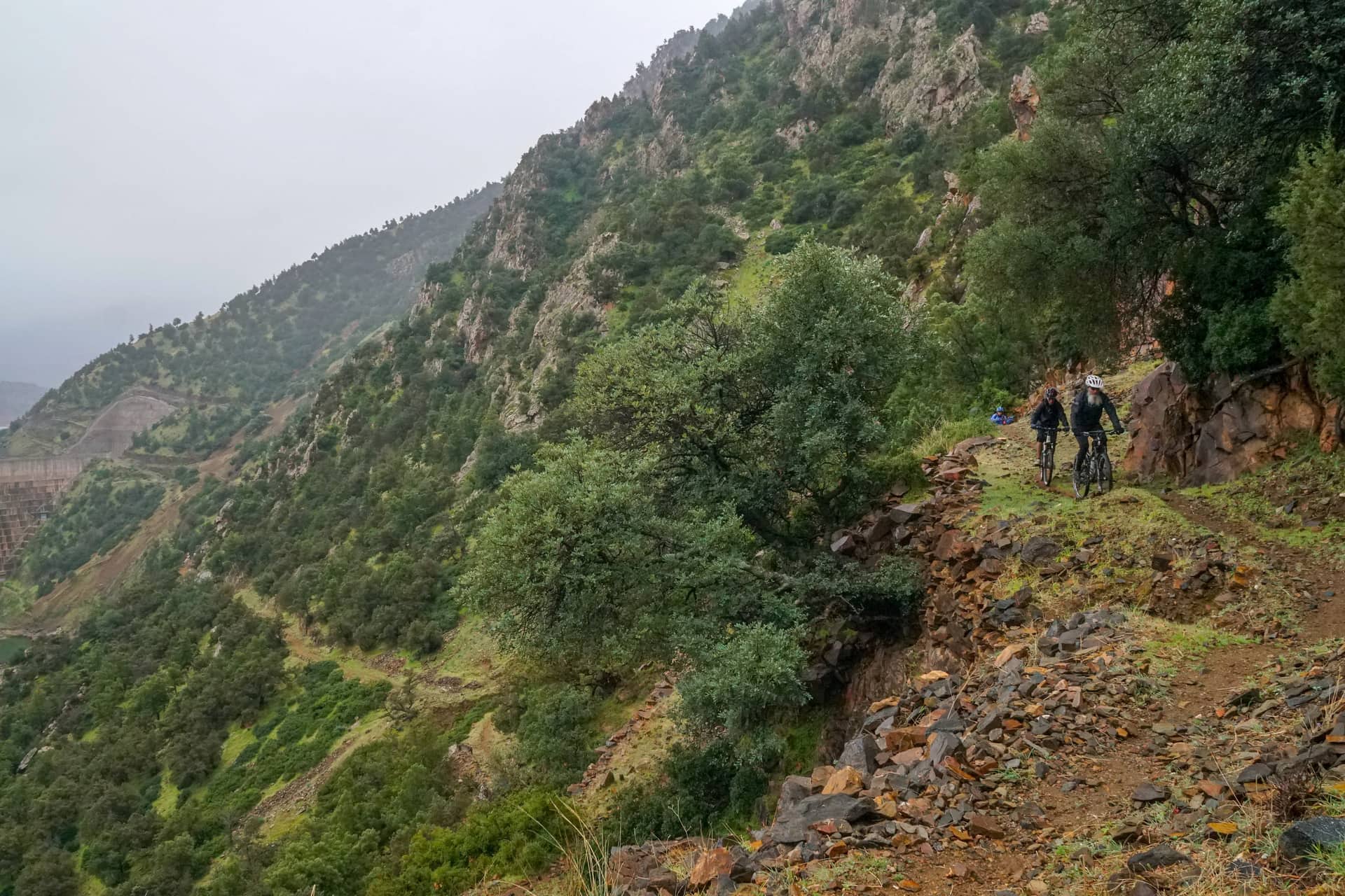 Two riders on a narrow trail traversing a steep green hillside above a dam