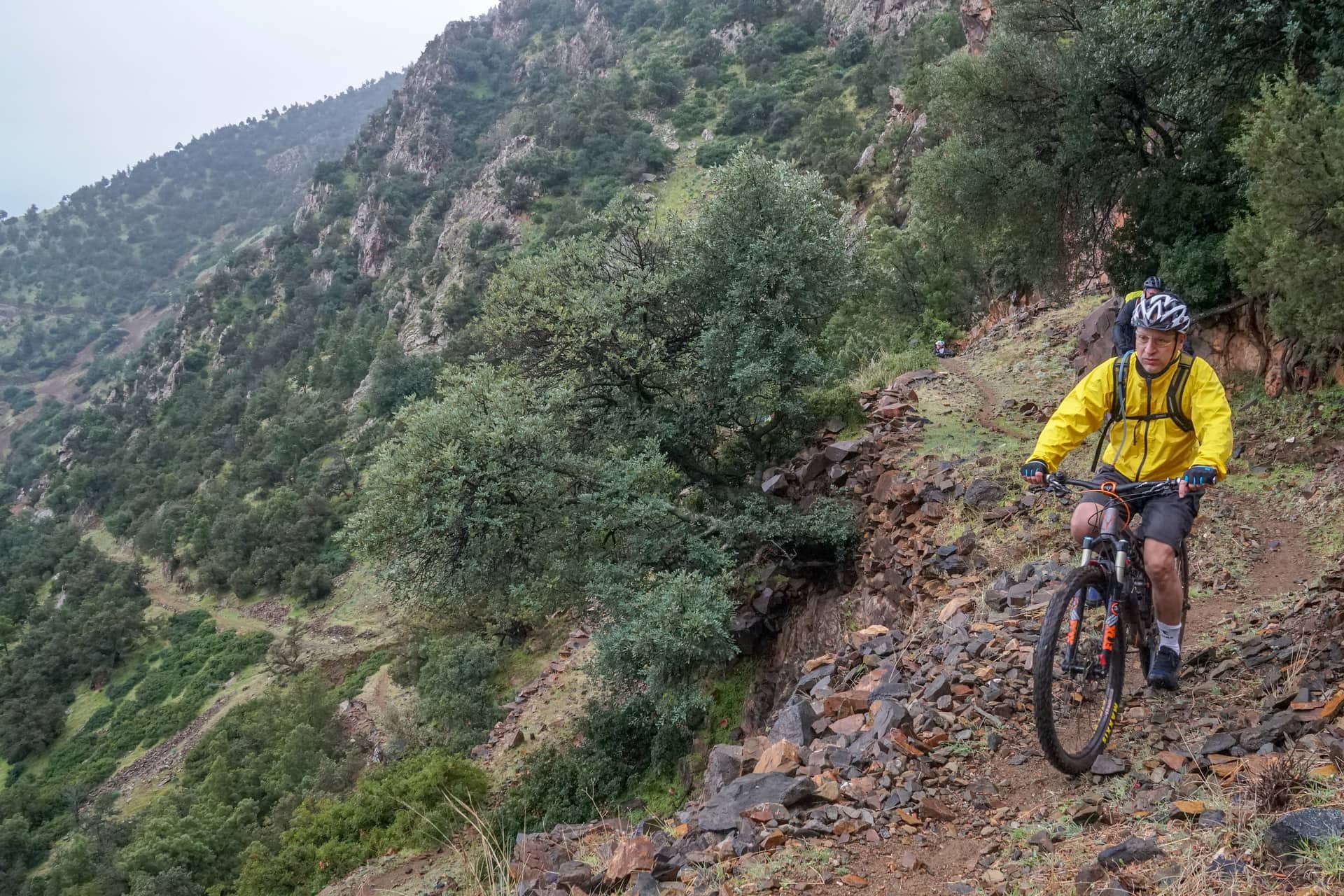 Mountain biker riding a rocky singletrack traverse high above a wooded Atlas valley