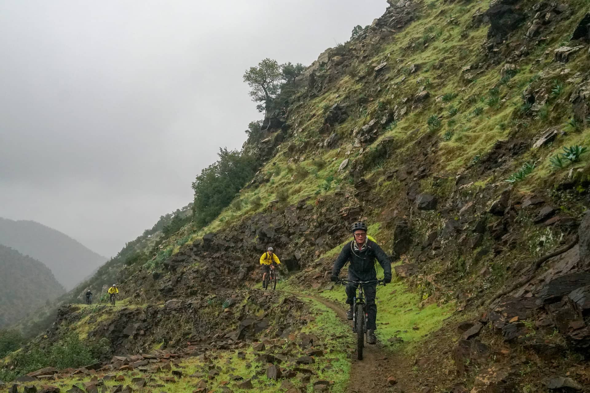 Two riders on a narrow trail traversing a steep bare mountainside in mist