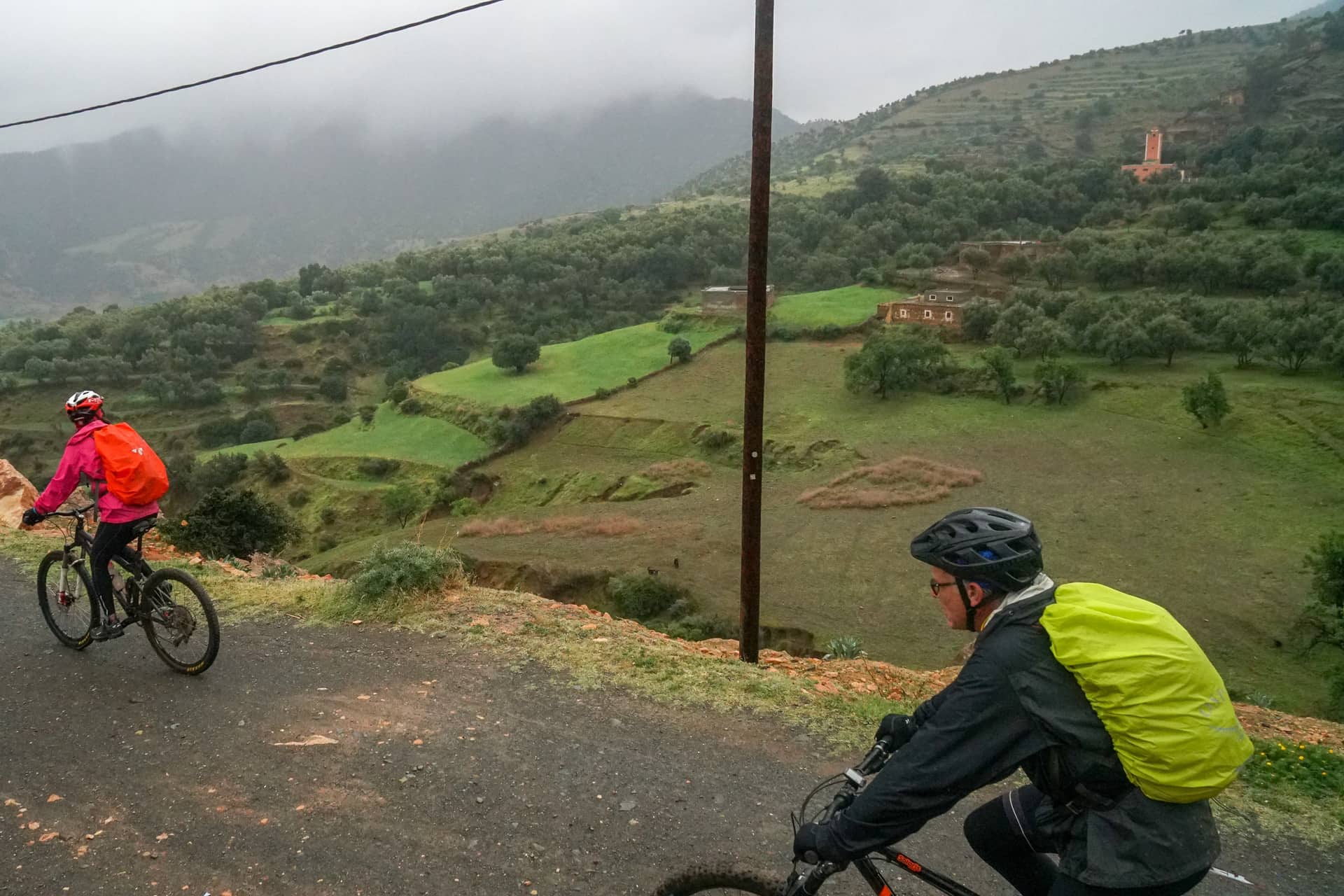 Two riders on a wet mountain road looking across terraced green fields