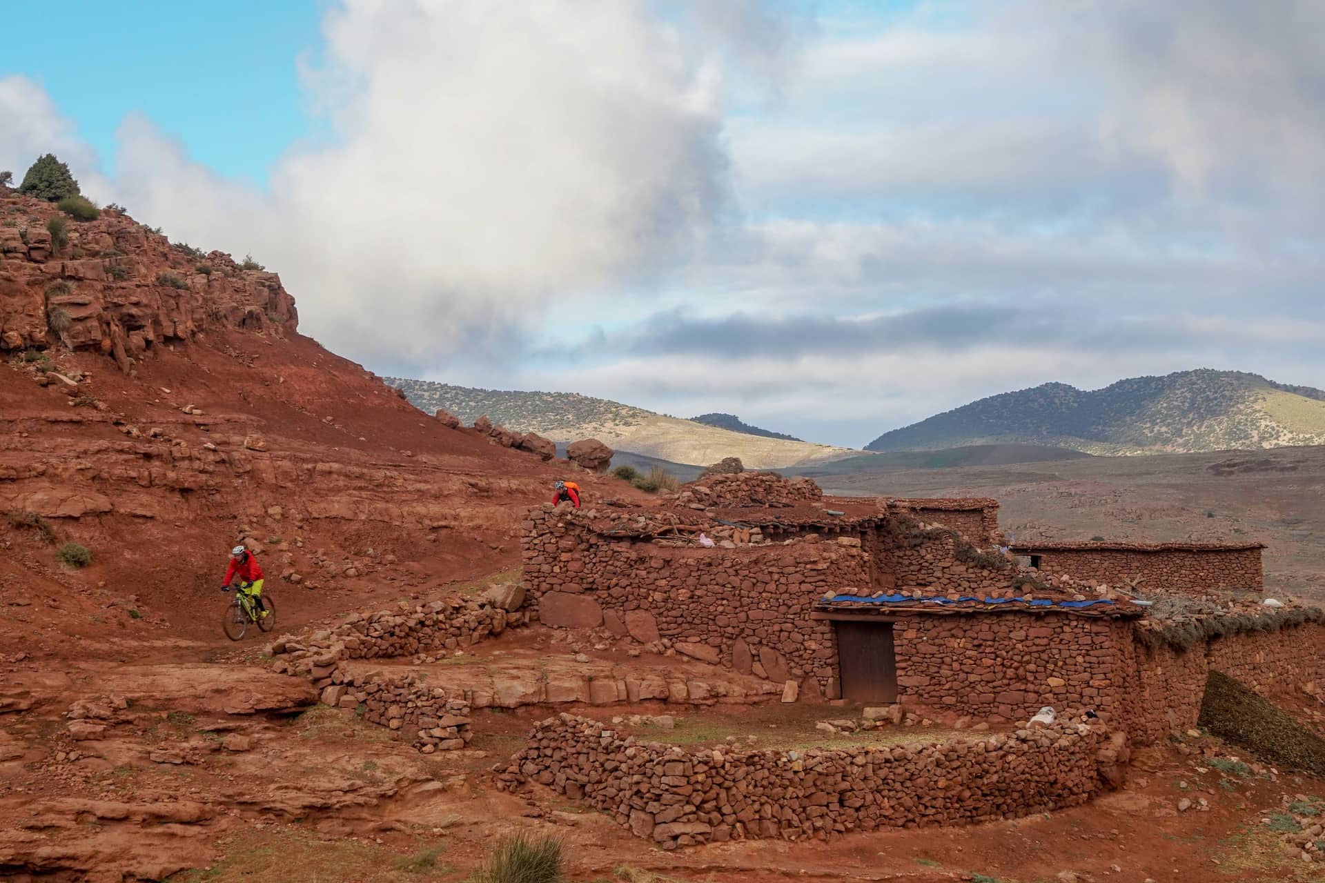 Two riders threading between red stone houses in a high Atlas village