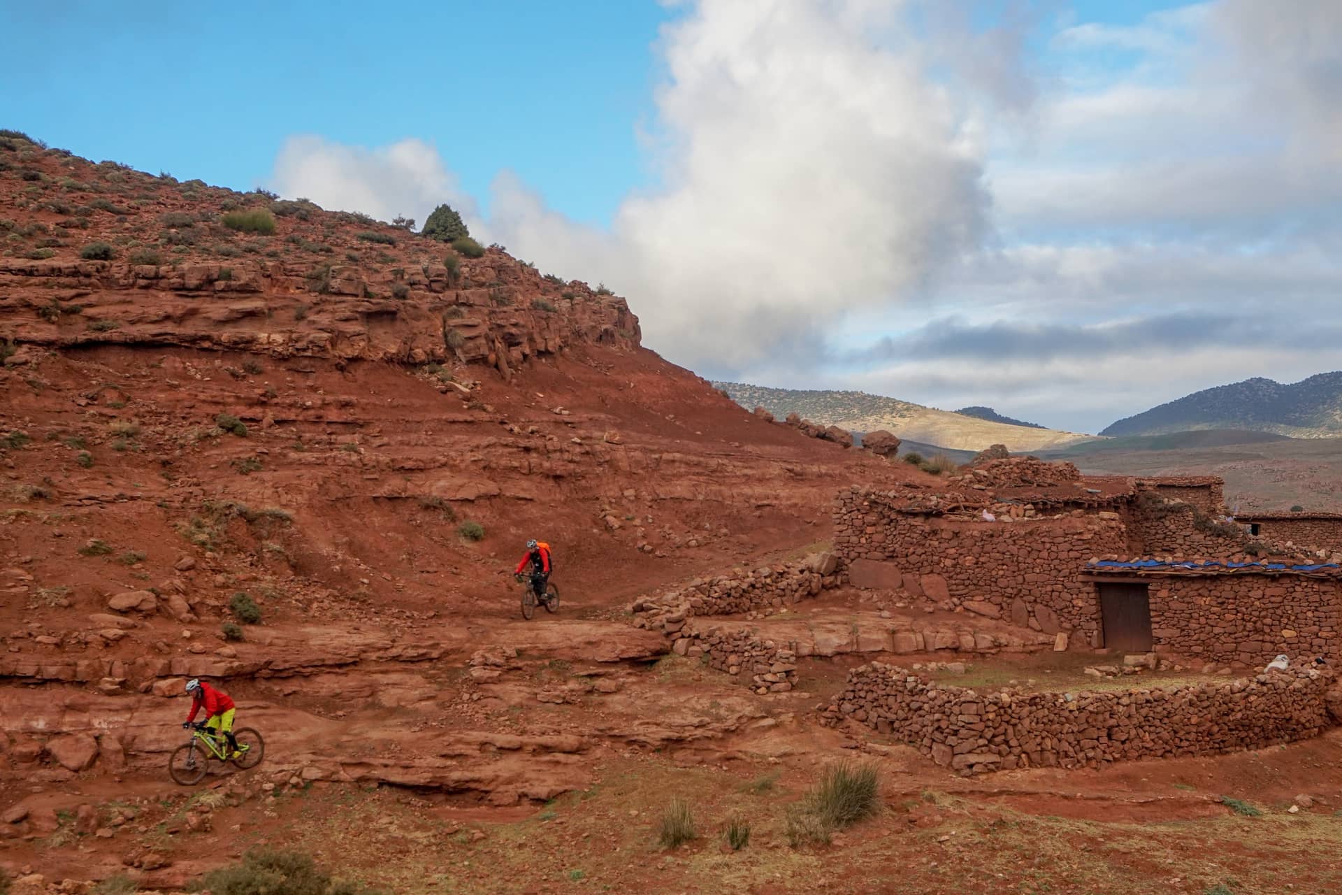 Two riders on red rock slabs beside a dry-stone Amazigh village