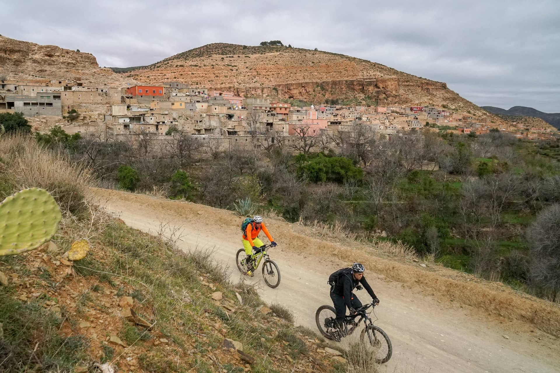 Two riders on a dirt road below an Amazigh village built into the hillside
