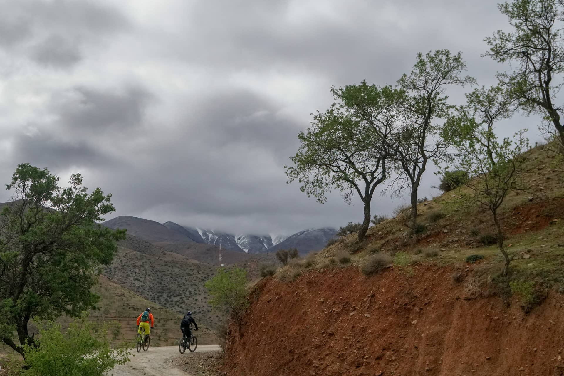 Two riders on a dirt road with snow-dusted peaks under heavy cloud ahead