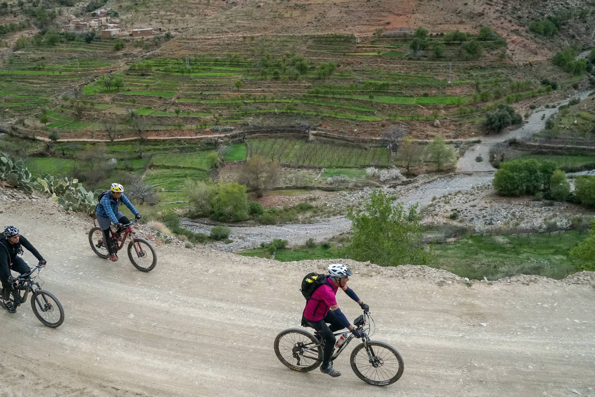 Three riders climbing a graded piste above terraced fields in the Atlas foothills