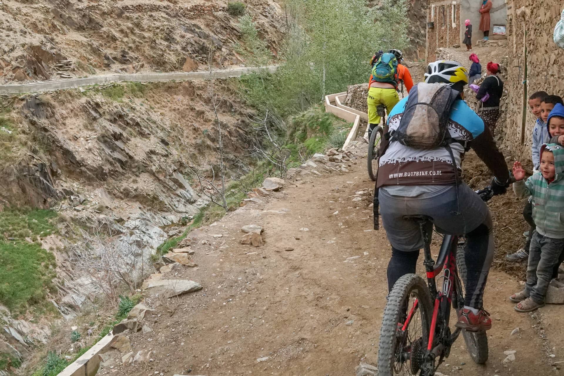 Riders passing through a village lane as children wave from the roadside