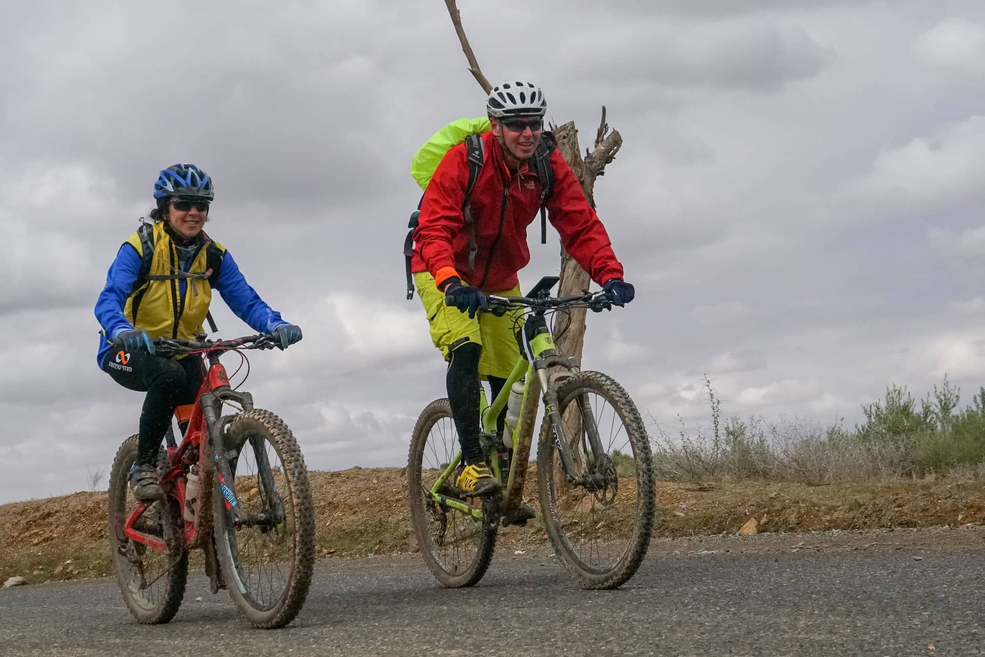 Two riders climbing a tarmac road on a grey day, smiling at the camera