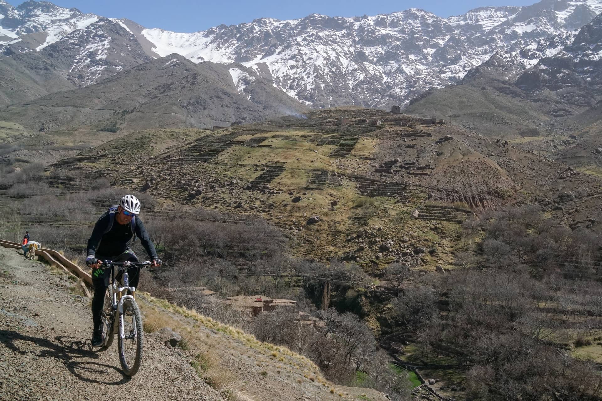 Rider climbing a piste with the snow-covered High Atlas and terraced fields behind
