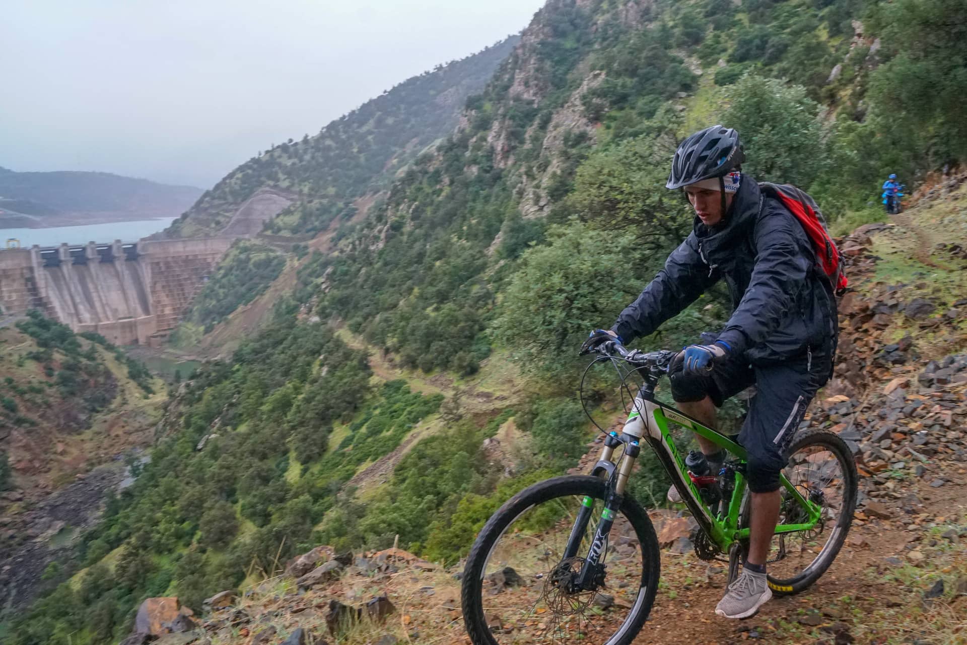 Rider on a rocky trail high above a reservoir, with the dam wall behind