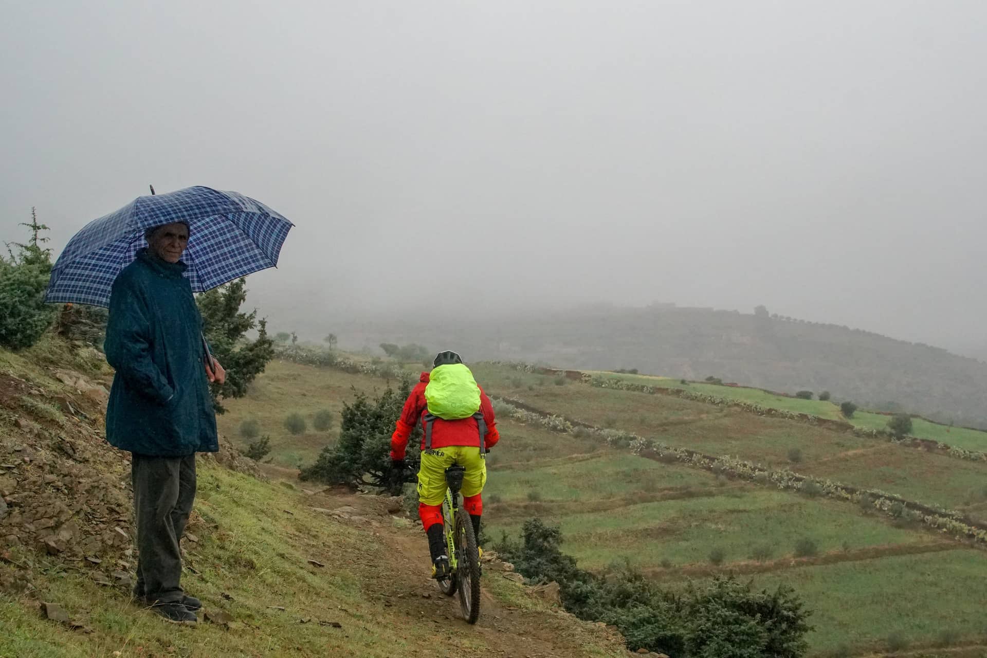 A villager sheltering under an umbrella watches a rider descend a wet trail